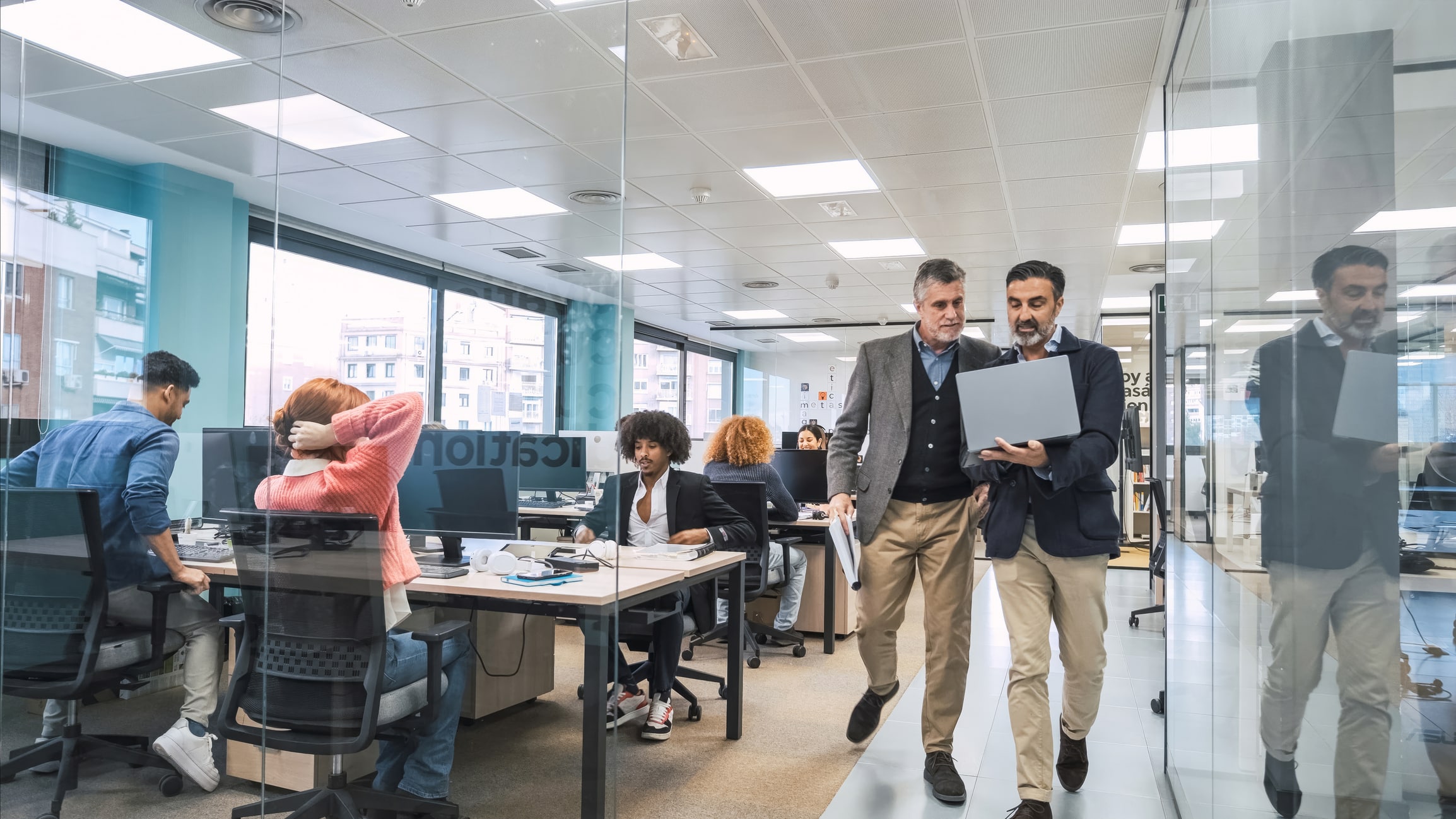 Two managers walking through an open plan office reviewing figures on a laptop
