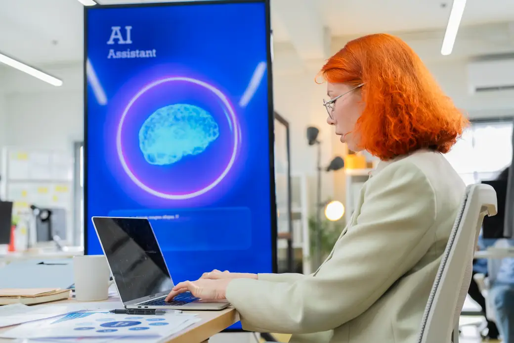 An employee working on a laptop beside a large screen showing an AI assistant