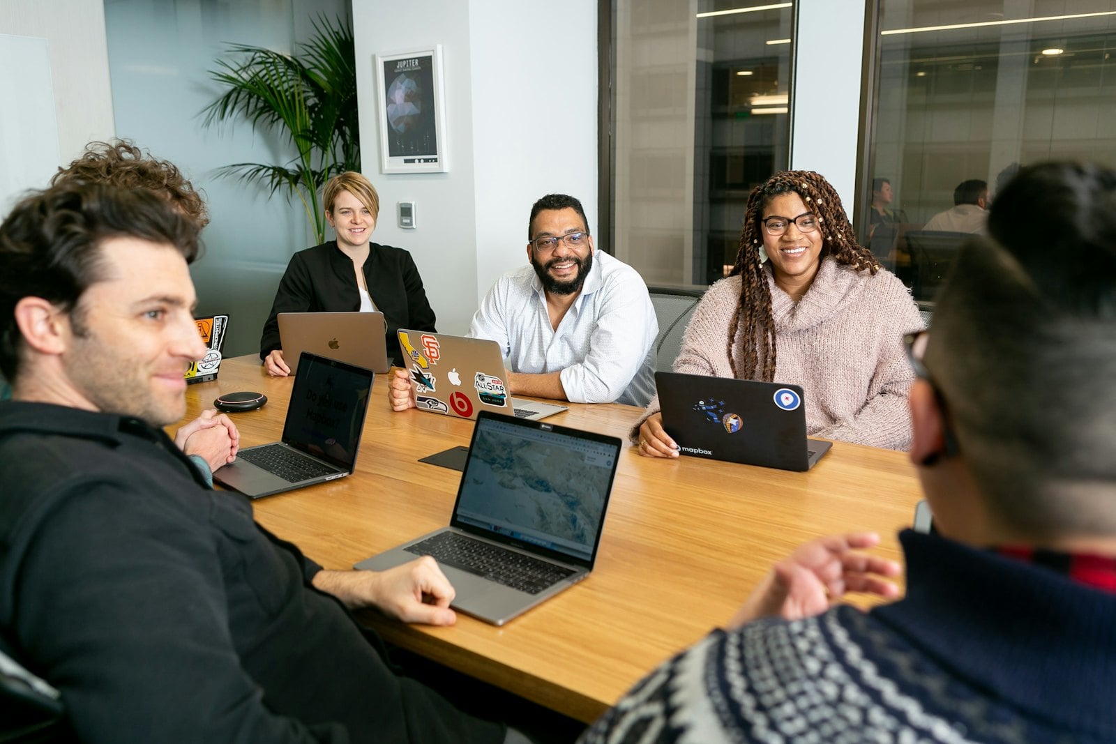 Four people around a boardroom table with laptops open, listening to a colleague
